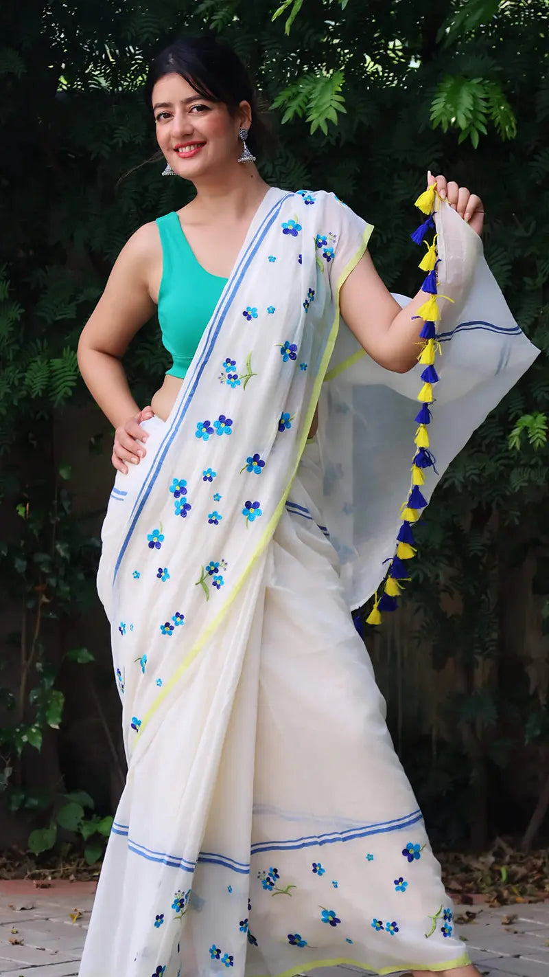 Woman in a white saree with blue and yellow tassels, standing outdoors.