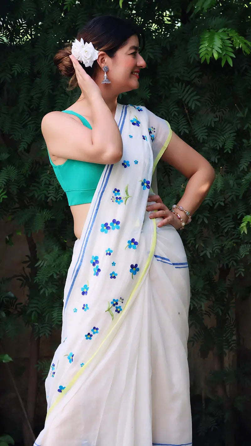 Woman wearing a white mul cotton saree with blue floral patterns and a green blouse, standing against a green leafy background.