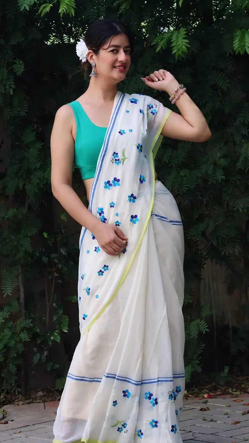 Woman wearing a white mul cotton saree with blue floral patterns and a green blouse, standing outdoors.