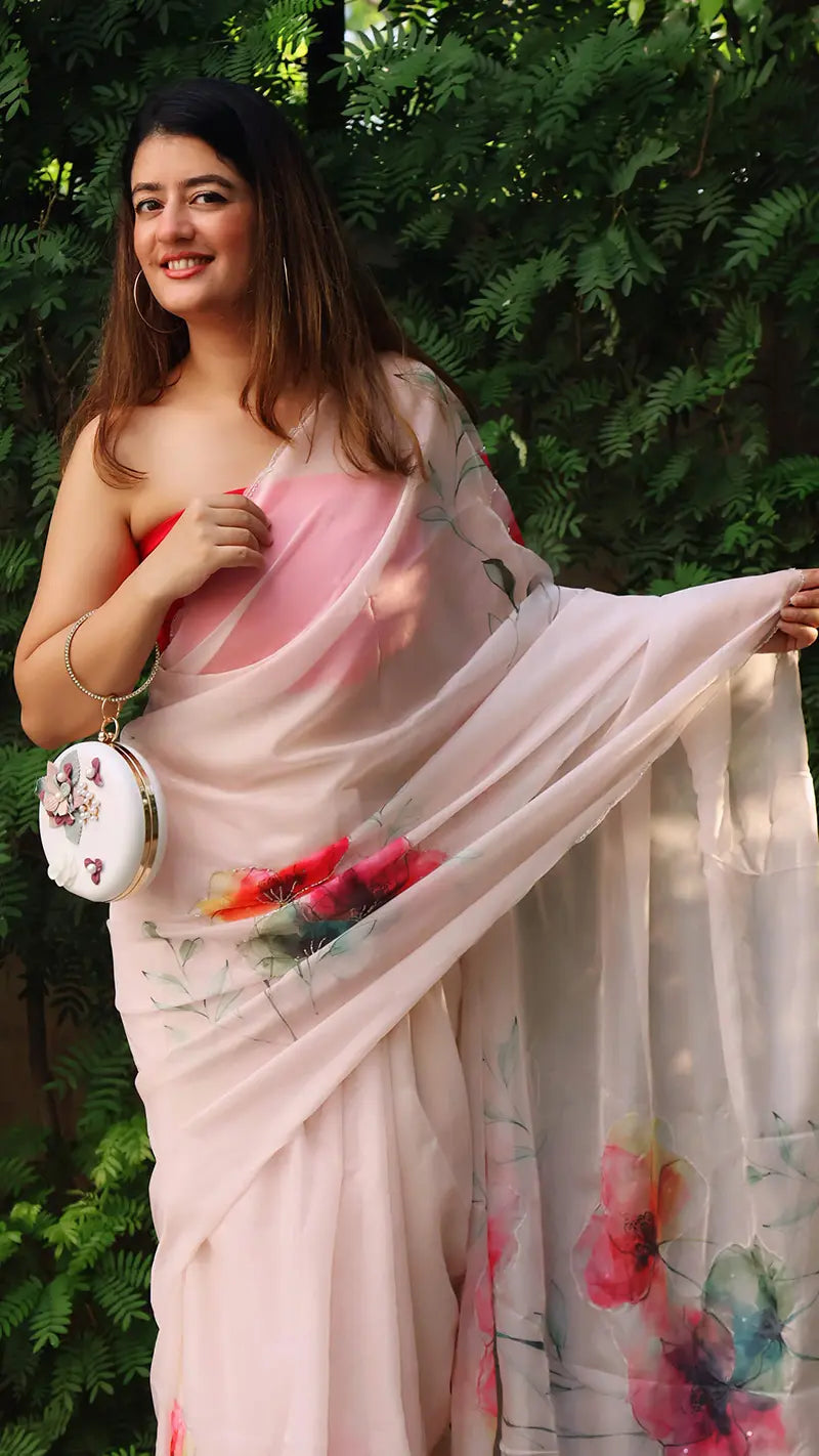 Woman wearing a light pink saree with floral patterns against a green foliage background